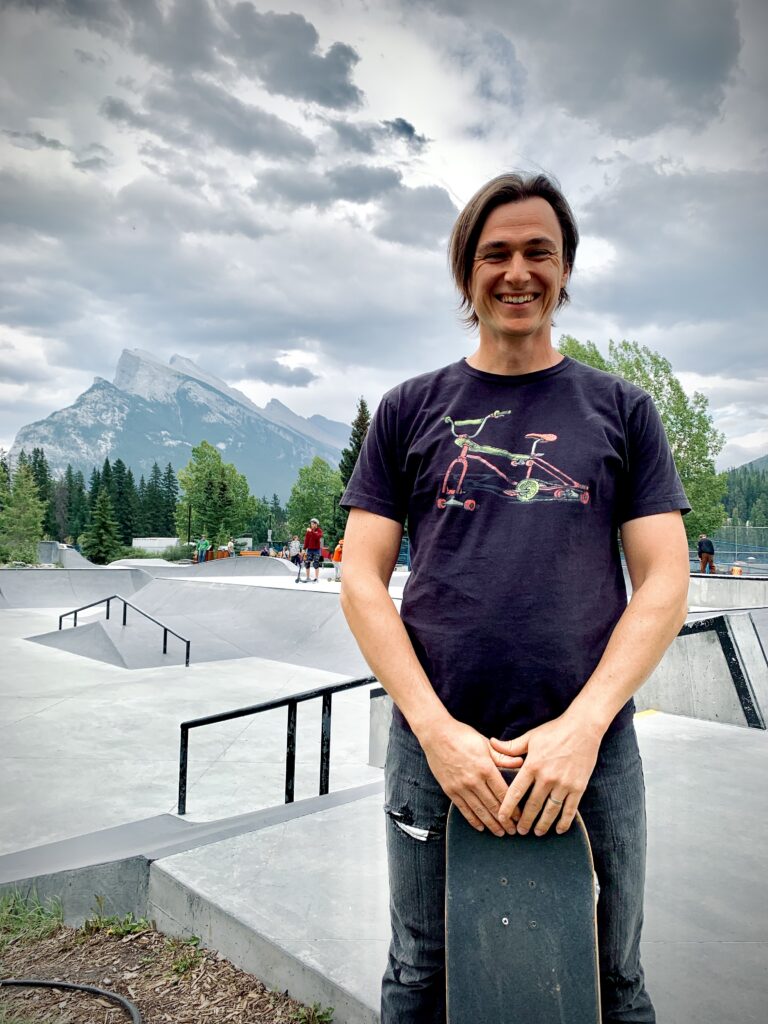 Kevin is standing in the forefront of the photo holding a skateboard. There is a skate park behind him and mountains in the background.